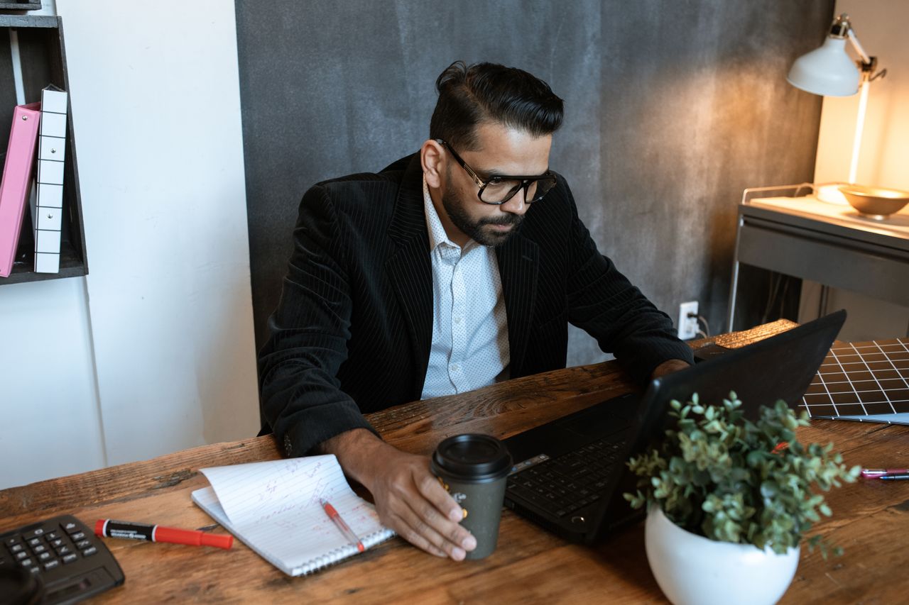 Man concentrating on laptop in a modern home office setting, coffee in hand.